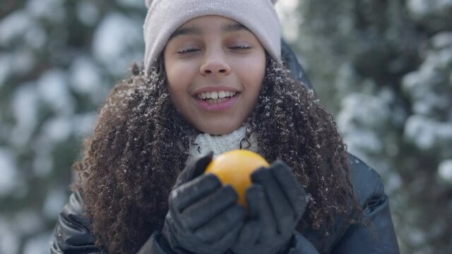 Close-up portrait of positive cute teenage girl smelling healthful vitamin orange standing in winter forest. Happy carefree African American teenager posing with fruit looking at camera smiling