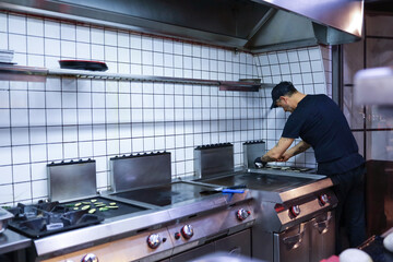 Young chef prepares grilled vegetables on cast iron griddle in his restaurant. Healthy food preparation concept.