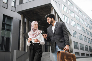 Arabian business colleagues in formal clothes walking on street and discussing some working issues. Young man with suitcase using smartphone, woman in hijab carrying clipboard.