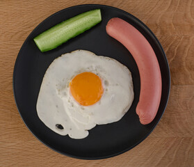 Fried egg, sausage and two pieces of cucumber lie on a black plate on a wooden board
