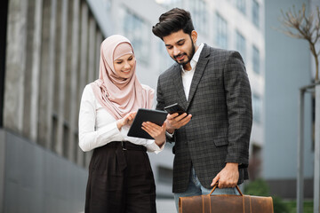 Charming woman in hijab with digital tablet and muslim man with smartphone standing together near office building. Two colleagues using modern gadgets for work outdoors. - Powered by Adobe