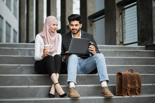Stylish muslim man and charming woman in hijab sitting together on stairs, smiling and talking. Two freelancers using laptop for for work outdoors.