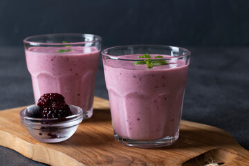 Homemade smoothies with frozen blackberries in two transparent glass on wooden board on dark gray background. Close-up