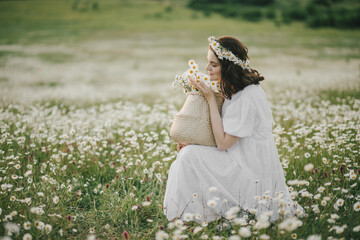 Young woman wearing white dress holding straw basket with flowers on chamomile field.