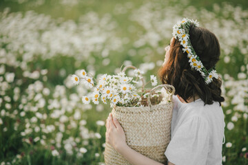 Young woman wearing white dress holding straw basket with flowers on chamomile field.