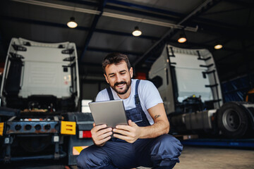 Smiling tattooed bearded blue collar worker in overalls using tablet to check on delivery while...