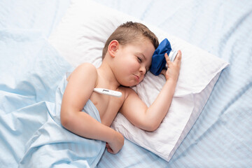 Sick little child boy with headache lies in his bed
and measures the temperature and cools the head with a cold pack