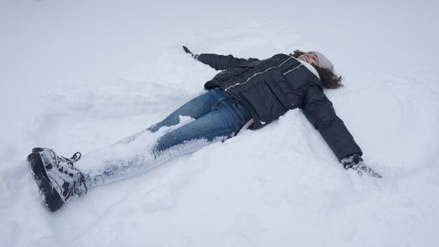 Wide Shot Of Carefree Happy African American Teen Girl Making Snow Angel Lying On Snowy Ground In Winter. Laughing Cheerful Teenager Enjoying Cold Weather Travelling Outdoors. Tourism And Leisure