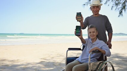 Asian senior couple tourist showing display digital vaccination passport certificate from smartphone at beach. Couple elderly senior retire resting and relax wife is disabled and sitting on wheelchair - Powered by Adobe