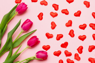 a group of red fresh tender tulips with green leaves lie on a pastel pink background with small hearts. Valentine's day concept. View from above