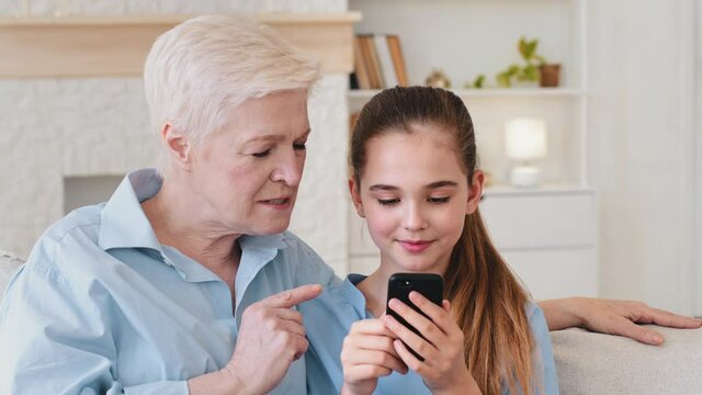 Adorable kid girl holding smartphone, looking funny pictures, video with smiling mature grandma relaxing together on couch. Little granddaughter playing mobile on cellphone games with granny at home
