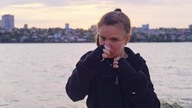 A young girl boxer strikes a series of punches in front of the camera against the background of a river in the city on the street. Sports, kickboxing. A moving frame.