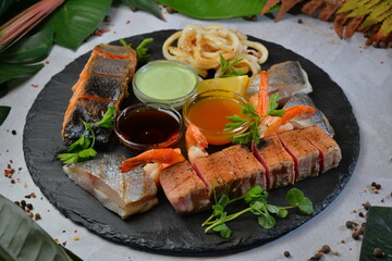 Assorted grilled fish fillets served with lemon slices and thyme on black round board - Image