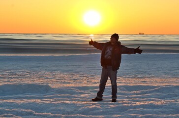 A man standing on the shore of a winter icy sea against the backdrop of a sunset