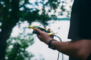 The guy is holding a portable charger with a smartphone in his hand. Man on a background of nature...
