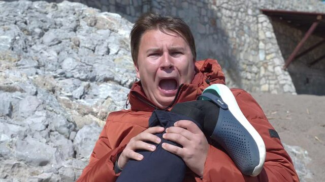 Young Caucasian man in down jacket sits with his leg in hands and screams in pain because he fell and broke leg in accident. Medical insurance. The actor overacts.