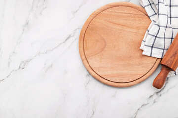 Empty wooden pizza platter with napkin and rolling pin set up on marble stone kitchen table. Pizza board and tablecloth on white marble background.