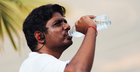 Fototapeta na wymiar Close up Asian young sport runner black man wear athlete headphones he drinking water from a bottle after running at the outdoor street health park, healthy exercise workout concept