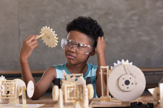 Happiness cheerful child curly black hair clever wearing safety glasses looking parth of  working model of a mechanism made of wood on table.Education and innovation concept.