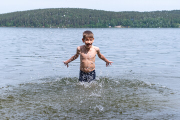 Fototapeta premium Cute happy boy makes water splash in the lake. The child bathes in the lake on a sunny day. Cheerful happy childhood concept