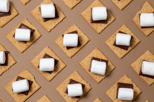 s'mores ingredients. graham cracker squares with chocolate, marshmallows on a brown background. ready to roast, geometric pattern