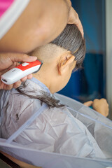 Asian child getting haircut at home from the father. Young boy save his head using electric razor.