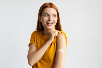 Covid-19 vaccinated caucasian smiling young woman showing arm with plaster, gray background - obrazy, fototapety, plakaty