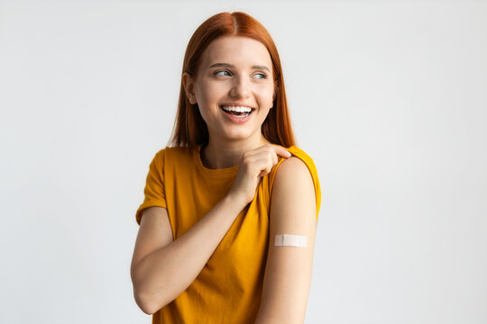 Covid-19 vaccinated caucasian smiling young woman showing arm with plaster, gray background