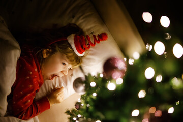 Little cute toddler girl in bed under Christmas tree and dreaming of Santa at home, indoors. Traditional Christian festival. Happy kid child waiting for gifts on xmas. Cozy soft light