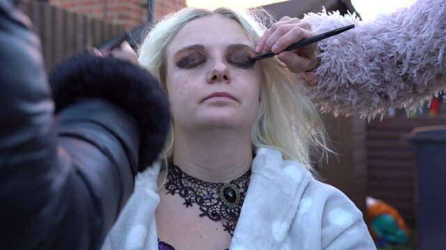 Pale blonde girl having dark make up applied for a photo shoot outside.