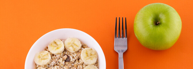 Fototapeta na wymiar muesli with banana on orange background with apple. High quality photomuesli with banana in a white plate on an orange background, a fork and a green apple . Healthy food and diet concept