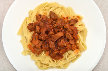 Close-up of Italian traditional tagliatelle pasta with tomato sauce, pork pieces, carrot slices, onion pieces and chopped parsley on the white plate. Top view.