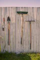 a rake and a shovel hang on a wooden white fence on a sunny summer day