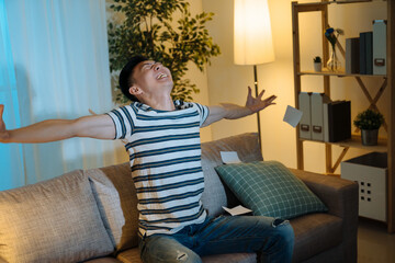 happy successful young Asian billionaire man smiling happily open arms stretching and looking up. Wealth investment economic concept. cheerful male sitting in home living room couch win lottery