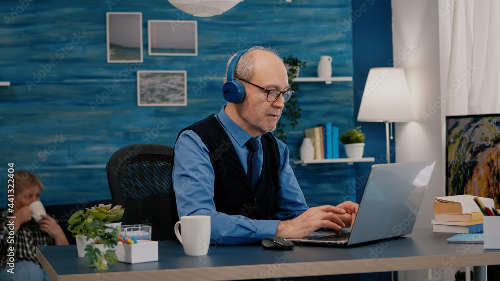Poster focused old entrepreneur with headphones listening music while checking graphics writing on laptop w