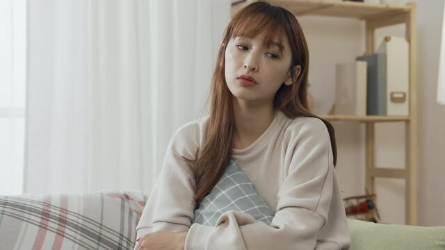 korean woman holding pillow in arms is thinking dully about her weekend plan in living room. boring asian young female sitting alone lets out a soft sigh feeling like doing nothing at home.