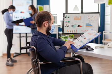 Handicapped entrepreneur with protective face mask working in new normal business financial company checking project documents, analysing data typing on pc sitting in wheelchair.