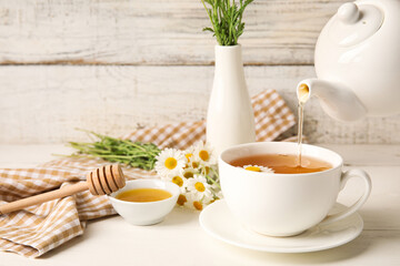 Pouring of tasty chamomile tea from teapot into cup on light wooden background