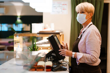 Mature woman wearing face mask working at cash register in cafe