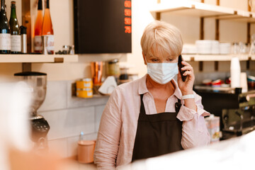 Mature woman wearing face mask talking on cellphone while working in cafe
