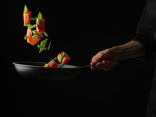 Red fish with herbs in a frying pan held by the hand of the cook. Black frying pan, black background. Focusing on the foreground. Levitation. Place for your insert.