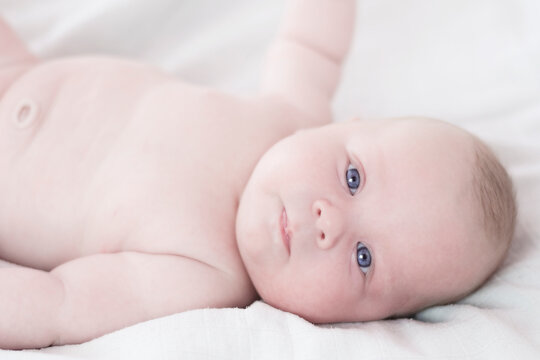 Caucasian infant lies on white blanket on his back and looks into the camera