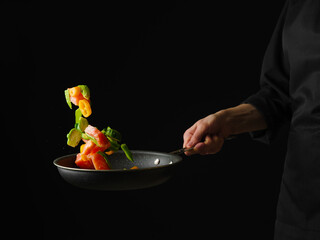 Pieces of trout with pieces of vegetables and herbs in a frying pan. Levitation. A chef in a black uniform holds a frying pan in his hand. There is an empty space for your signature.