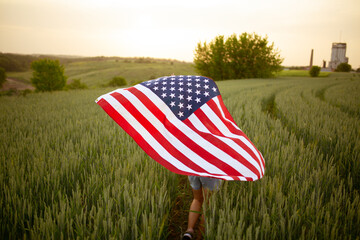 young happy beautiful  girl in pink top and jeans shorts with national American flag  in green wheat field at sunset.4th of July celebrating, view from back