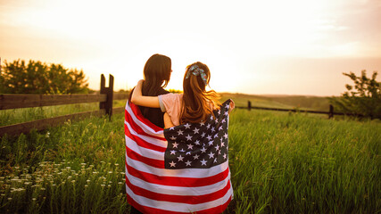 lesbian couple hugging each other cowering by USA flag view from the back countryside view in green wheat field at sunset. 4th of July celebrating, view from back