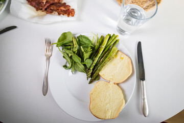 A hamburger bun on a plate with asparagus and a sife of bacon waiting for a hamburger.