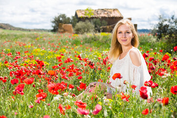 Naklejka na ściany i meble Beautiful young female in white dress in poppy field of wild flowers outdoor