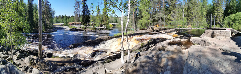A picturesque panorama of the Akhvenkoski waterfall on the Tokhmayoki River in Karelia with a suspension bridge over it on a clear summer morning.