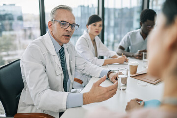 Portrait of white haired senior doctor talking to colleagues at meeting table during medical...