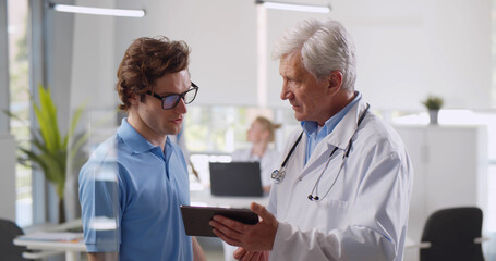 Mature male doctor pointing at digital tablet screen sharing health tests results to patient